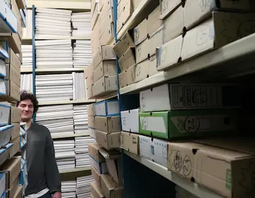 Aleksander, étudiant-chercheur, dans la salle de dépôt des archives face à nos 24 boites sur deux étagères de la salle de dépôt. Elles seront reconditionnées avant de rejoindre la salle des archives.